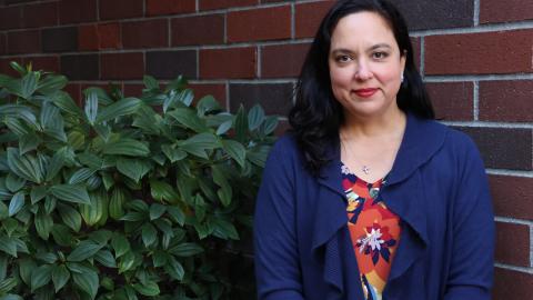 A woman with dark hair and a blue jacket stands against a brick wall next to some greenery.