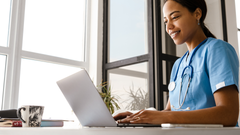 A healthcare professional in scrubs and a stethoscope uses a laptop at a desk with windows in the background.