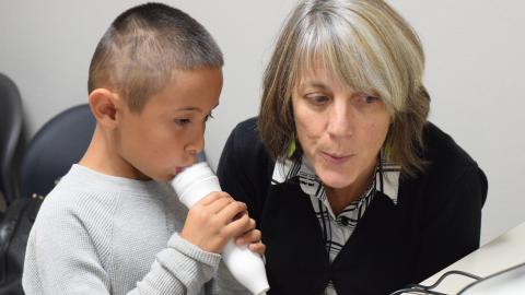 A young boy in a gray sweater breathes into a device measuring lung function, and a woman next to him in a black sweater demonstrates how to breathe while checking a computer monitor.