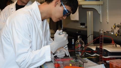 Man pipetting in lab, wearing lab coat and safety glasses.