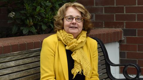 A woman wearing a yellow scarf and jackets sits on a bench looking at the camera.