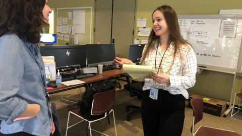 A woman in a white shirt talks with another woman in a blue shirt while standing in front of desks with laptops.