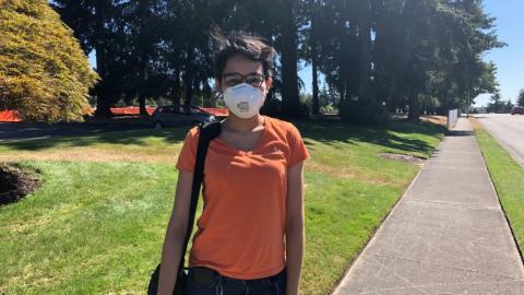 Pandher, wearing a face mask, standing next to a sidewalk with evergreens in the background.