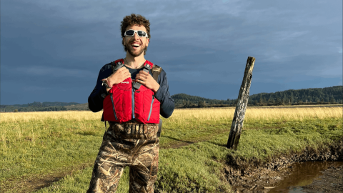 Orhan Eribac stands wearing a life vest by the water's edge in a field with mountains in the background.