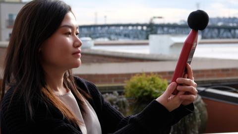 Shirley Huang stands on the roof of a building holding up a meter to take noise measurements, with a bridge in the background.