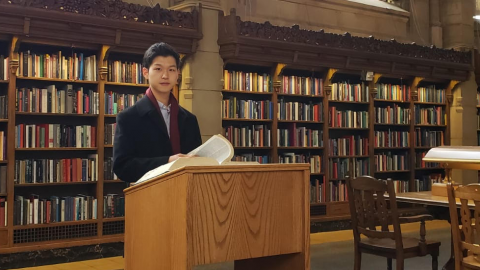 A man in a blue coat stands at a wooden lectern in a library.