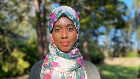 A woman in a floral head scarf smiling against a backdrop of trees.