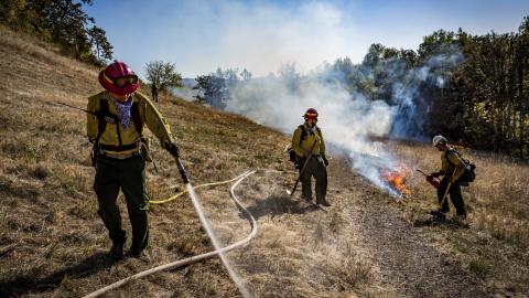 Three people in hard hats work on a hillside with a small fire burning, one spraying water from a hose.